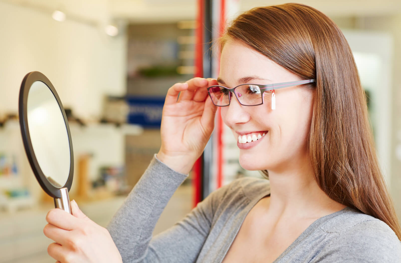 A girl trying on new glasses and examining herself in a small hand-held mirror.