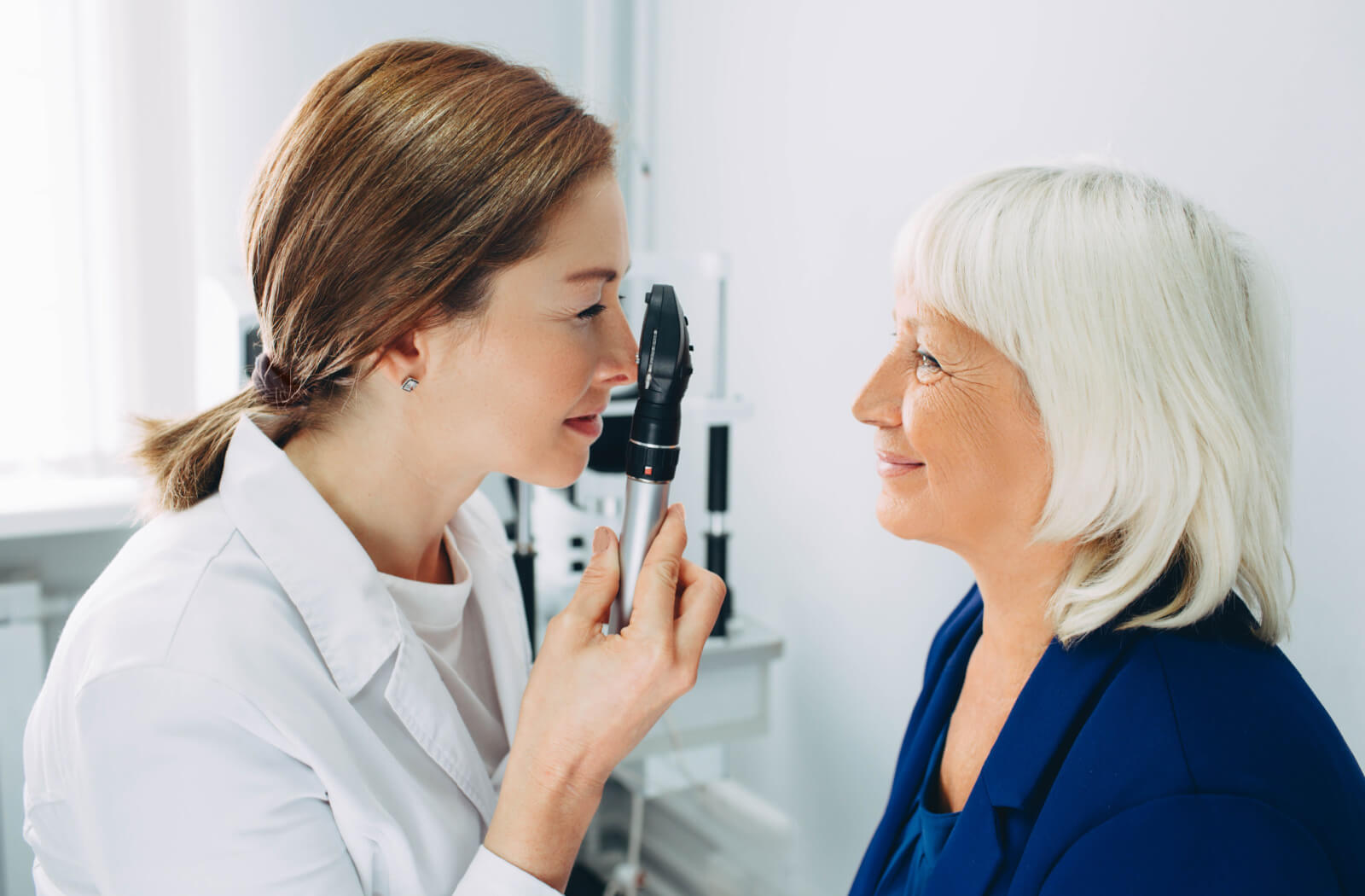 A woman optometrist is employing an ophthalmoscope while conducting an eye examination for an elderly woman