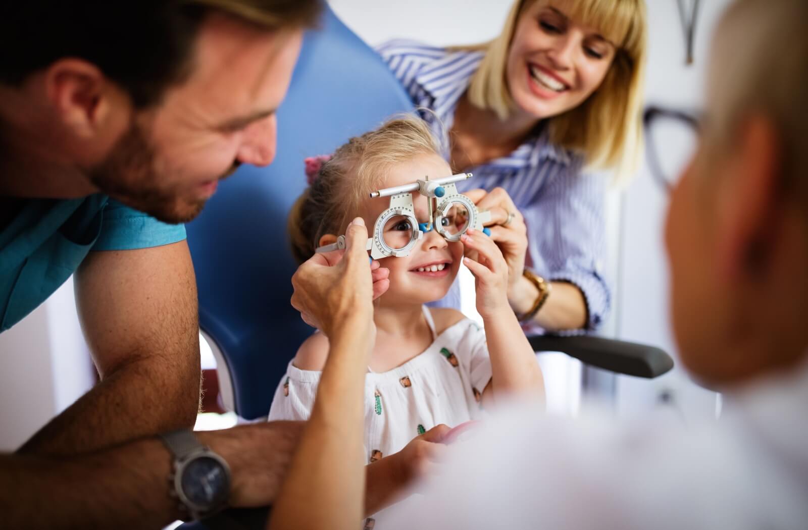 A young girl smiles as she gets her eyes checked by an optometrist. Her parents are on either side, smiling and supportive