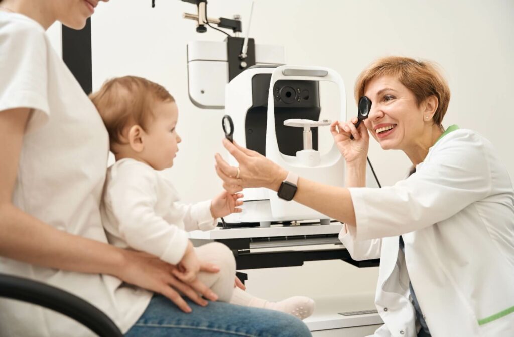 An infant on their mother’s lap receives their first eye exam from a friendly, smiling optometrist in a clean, white office