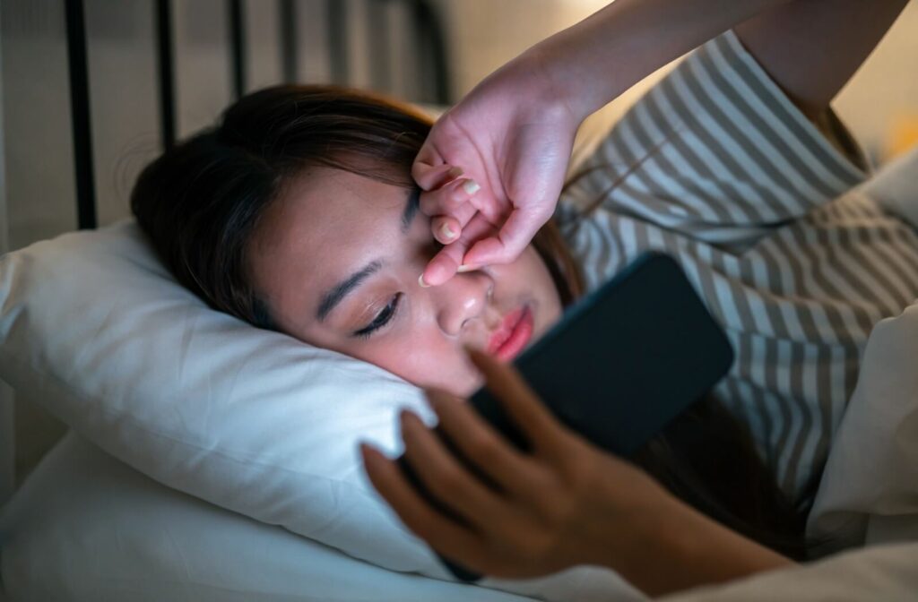 An image of a person laying in bed at night, while looking at their mobile device and rubbing their dry, irritated eyes.