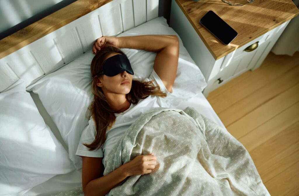 An image of a person sleeping in bed while using an eye mask to fully cover their eyes at night in order to prevent dry eyes.
