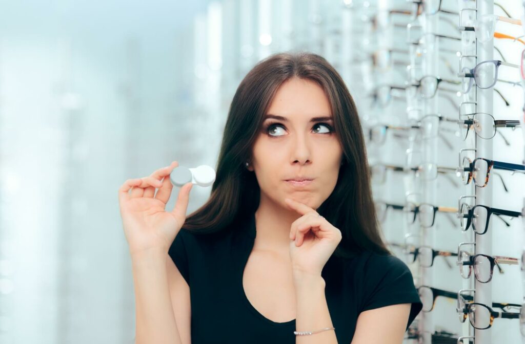 An adult pondering while holding a contact lens case next to a wall of glasses in a bright optometry clinic