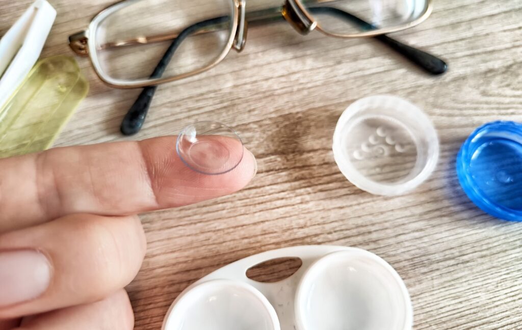 A person holds a contact lens up from their case. A pair of eyeglasses sits on the table below as a comfortable alternative to contact lenses.