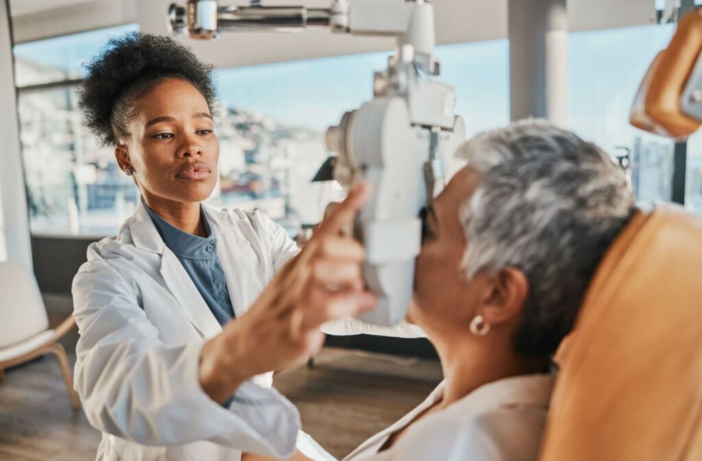 An eye care professional adjusting a diagnostic device during an eye exam for a seated patient in a clinic setting.