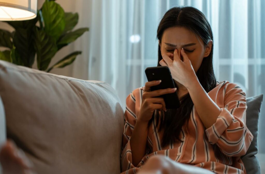 Person sitting on a couch, holding a smartphone and rubbing their eyes, appearing uncomfortable or fatigued.