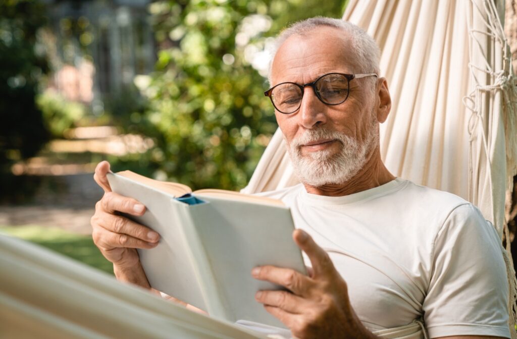 A senior man wearing glasses sits in a hammock reading a book
