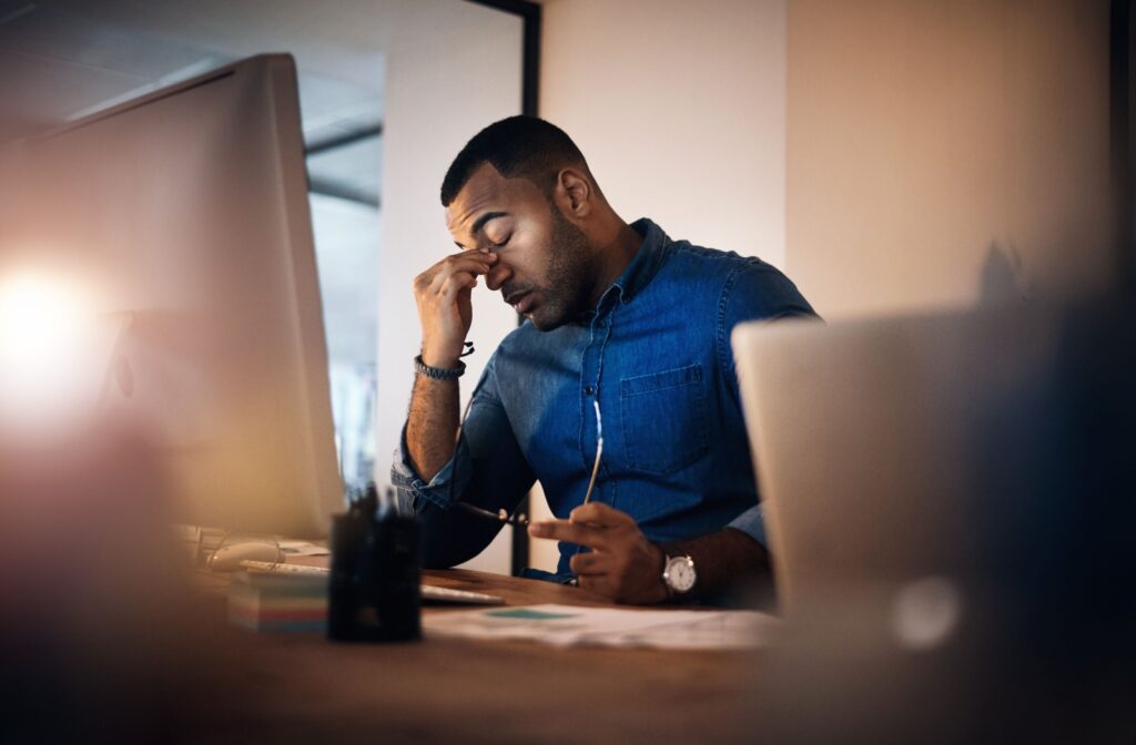 A person rubbing their eyes while sitting in front of the computer and holding a pair of glasses