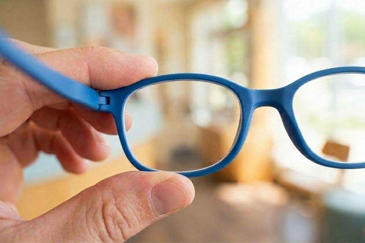 Hand holding blue-framed eyeglasses up to view, with a blurred indoor room visible in the background.